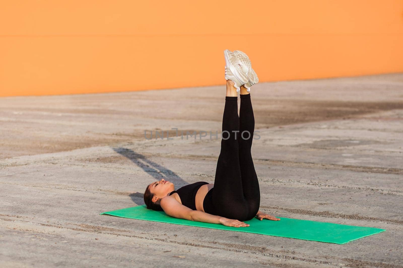 Fit slim young woman in tight sportswear, black pants and top, lying on mat keeping straight legs raised, training abdominal muscles, flexibility. Health care and weight loss, sport activity outdoor