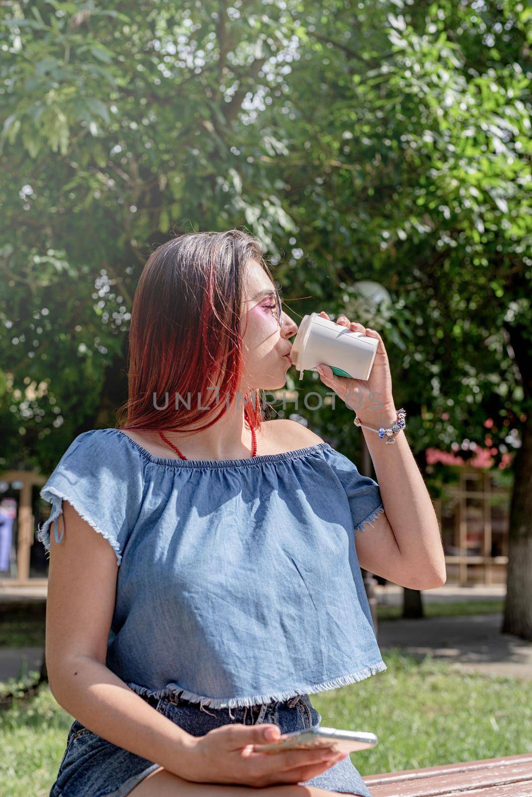 Attractive young woman in summer clothes and sunglasses holding cup of coffee in her hands and using smartphone videocall while sitting on bench in the park