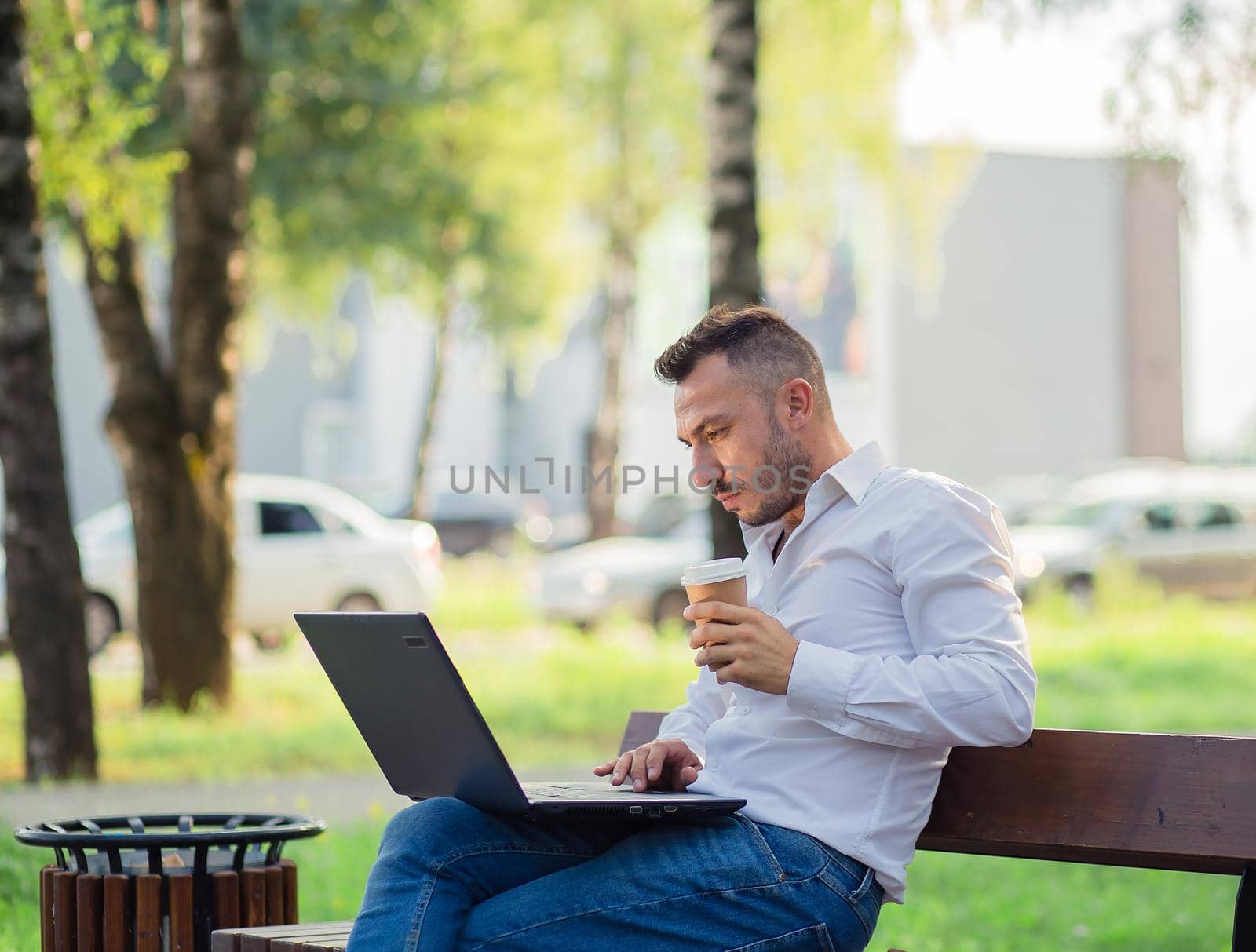 Serious businessman is working in the park with a laptop, drinking coffee. by anarni33