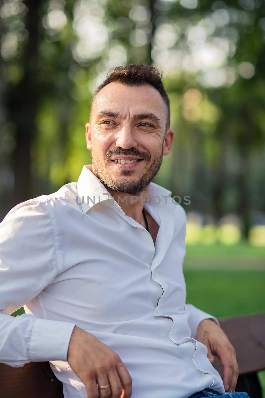 Smiling handsome man in a white shirt is resting sitting on a bench. Young man in city park, warm sunny summer day. Warm soft light, close-up.