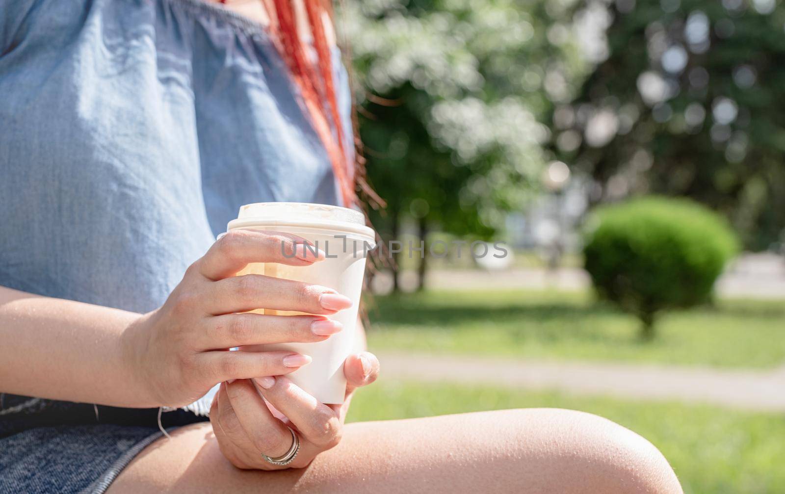 Attractive young woman in summer clothes and sunglasses holding cup of coffee in her hands while sitting on bench in the park