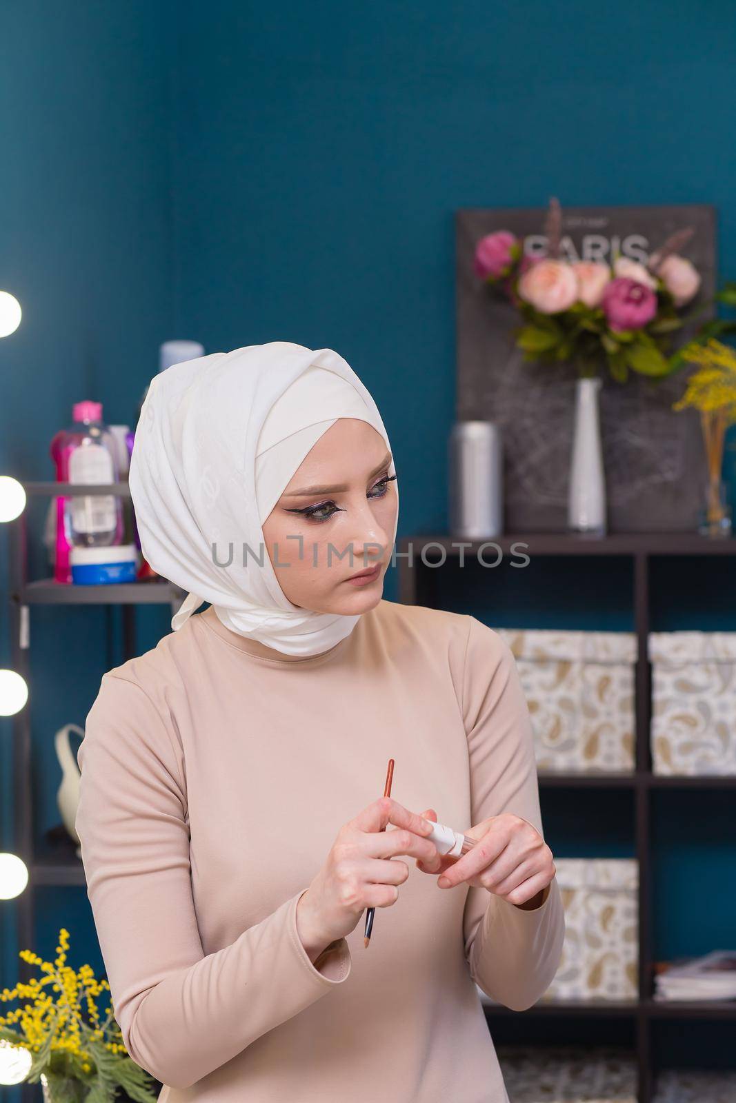 The makeup artist in a white scarf with a brush and mascara carefully smudges. Female master makes makeup to a young woman. Business concept - beauty salon, facial skin care, cosmetology.