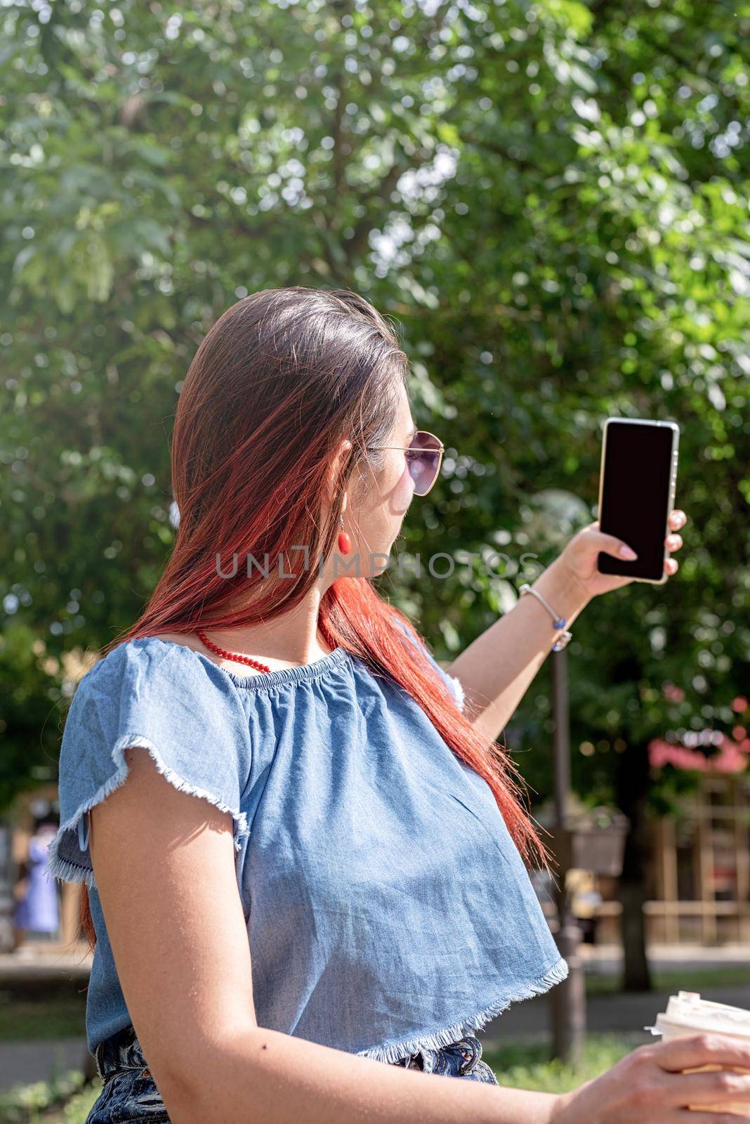 cheerful trendy woman with red hair drinking coffee at park, taking selfie, mockup screen by Desperada