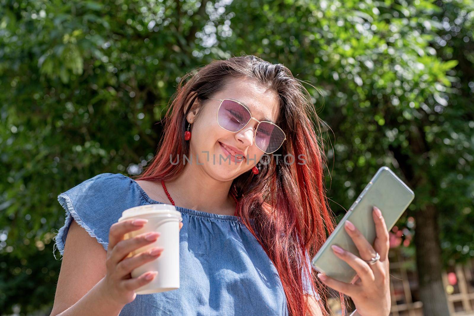Attractive young woman in summer clothes and sunglasses holding cup of coffee in her hands,using smartphone videocall while sitting on bench in the park