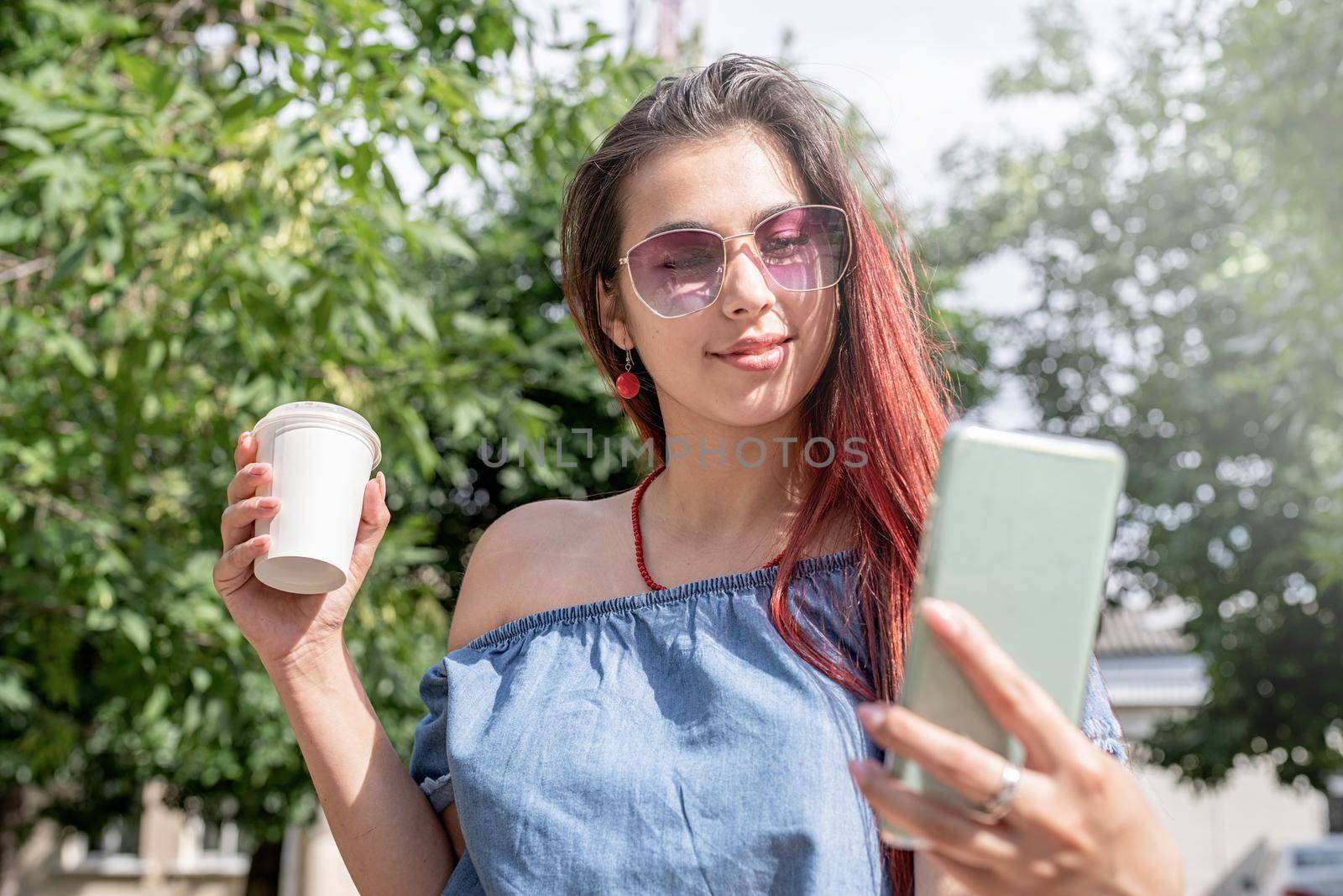 cheerful trendy woman with red hair drinking coffee at park, taking selfie by Desperada