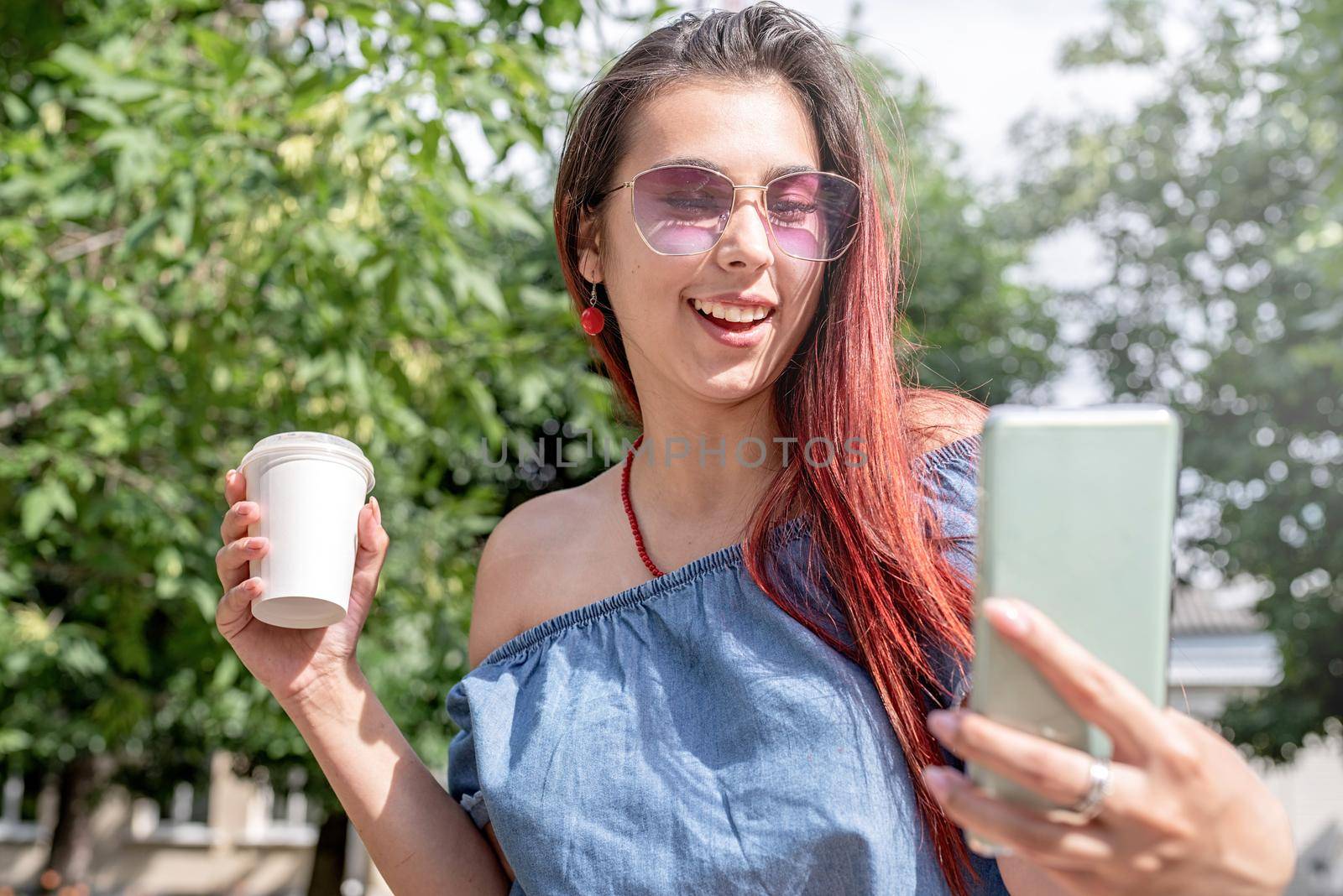 Attractive young woman in summer clothes and sunglasses holding cup of coffee in her hands,using smartphone videocall while sitting on bench in the park