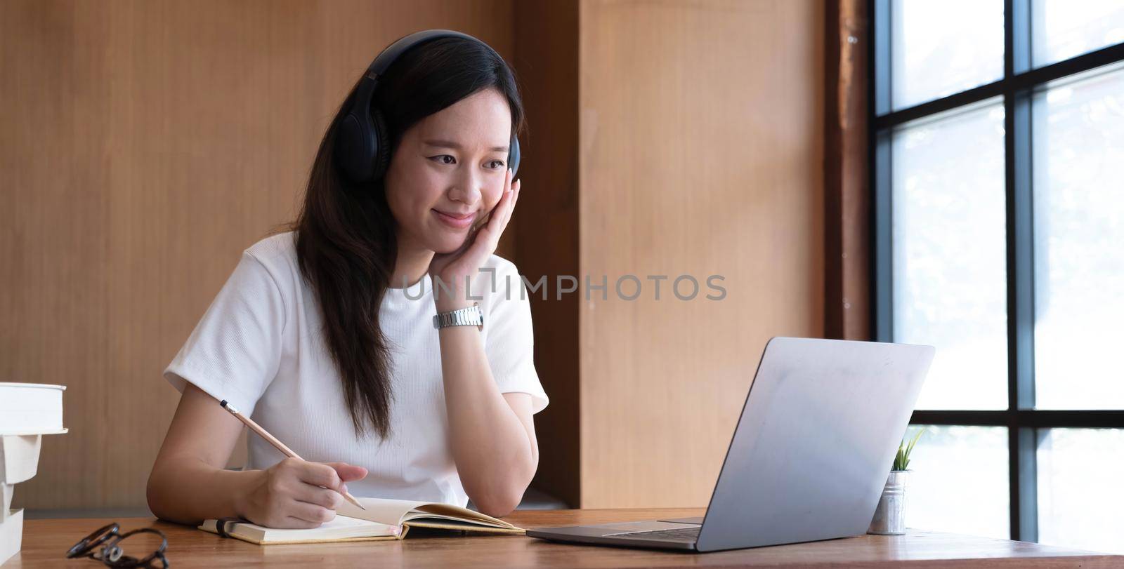 Smiling girl student wear wireless headphone study online with teacher, happy young asian woman learn language listen lecture watch webinar write notes look at laptop sit in cafe, distant education.