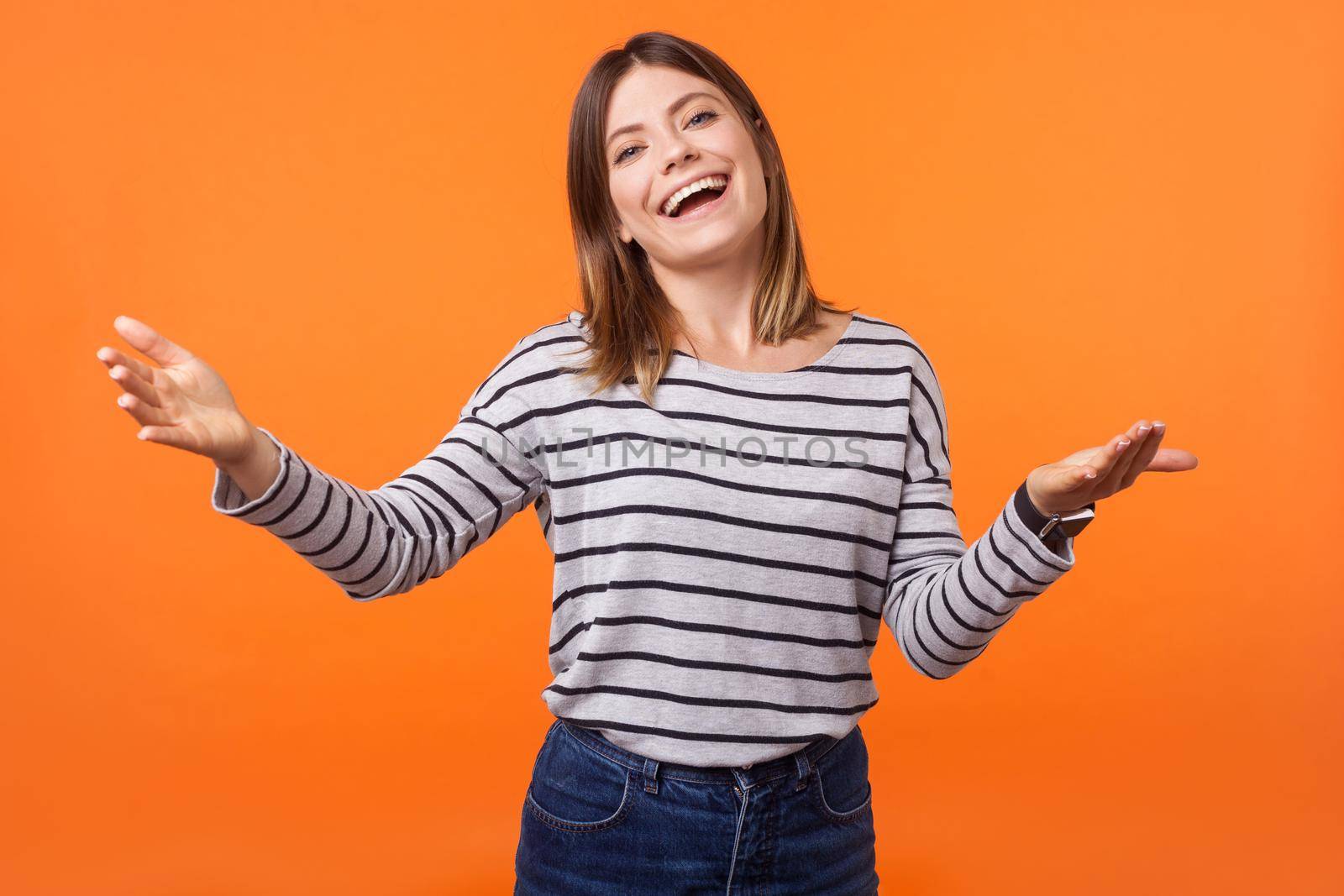 Free hugs. Come here. Portrait of friendly young woman with brown hair in long sleeve shirt standing with raised arms, welcoming and going to embrace. indoor studio shot isolated on orange background