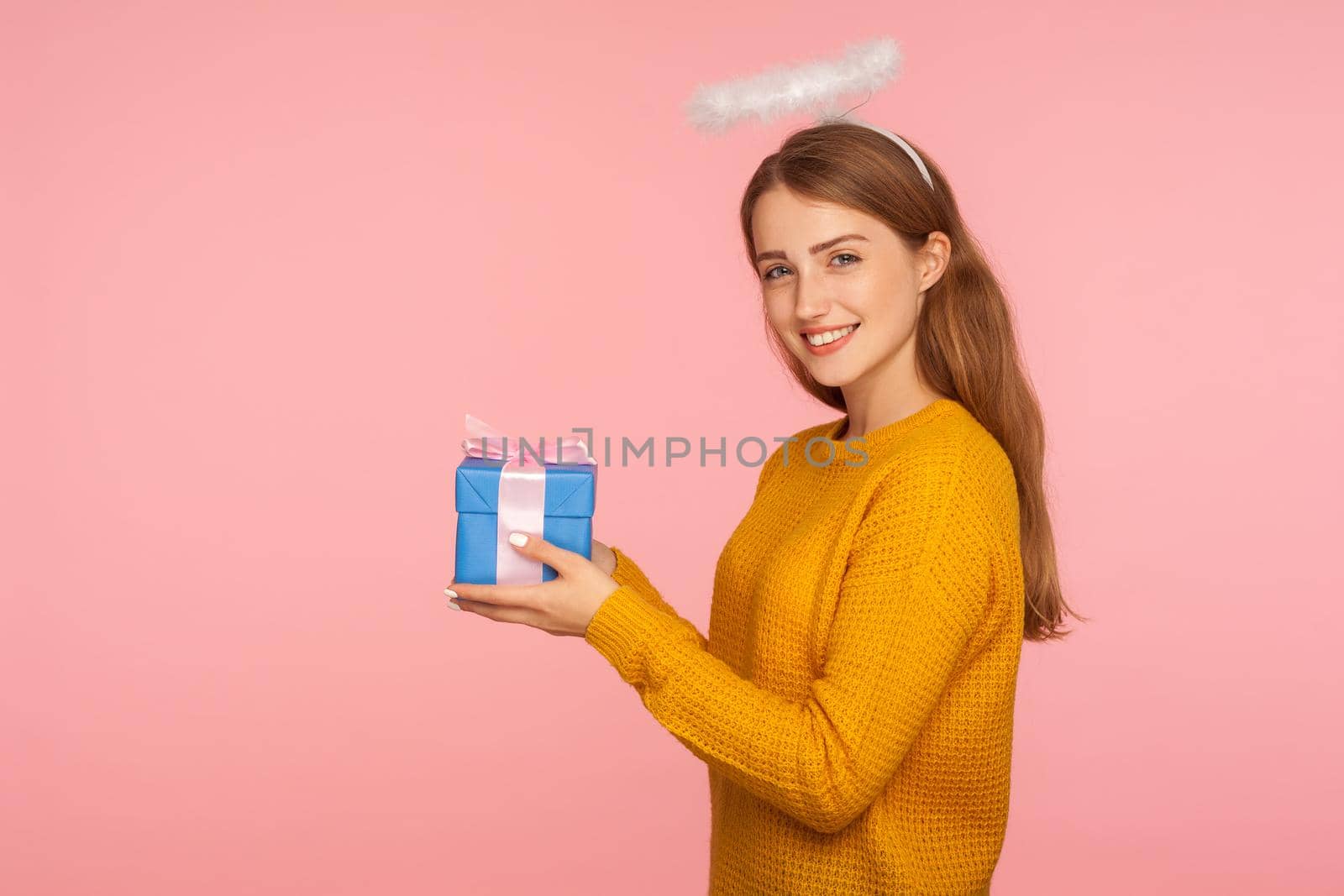 Charity and human kindness. Side view of angelic ginger girl with halo over head holding gift box and smiling at camera, giving holiday present to copy space. studio shot isolated on pink background