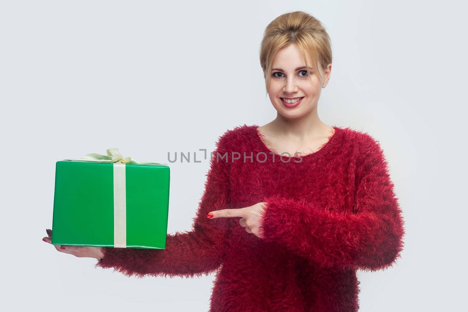 Portrait of happy young woman in red blouse standing and holding green gift box and pointing finger, looking at camera. Indoor, isolated, studio shot, copy space, gray background