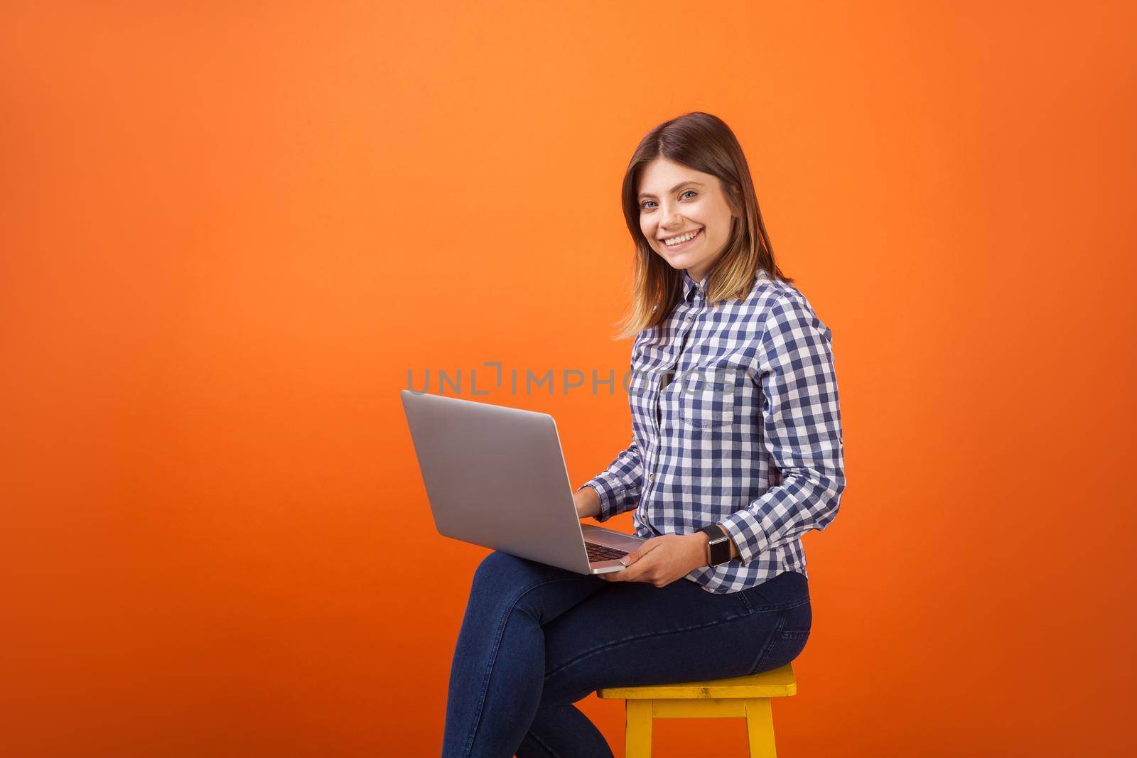Portrait of positive young woman with brown hair in checkered casual shirt and denim sitting with toothy smile, working on laptop, looking at camera. indoor studio shot isolated on orange background