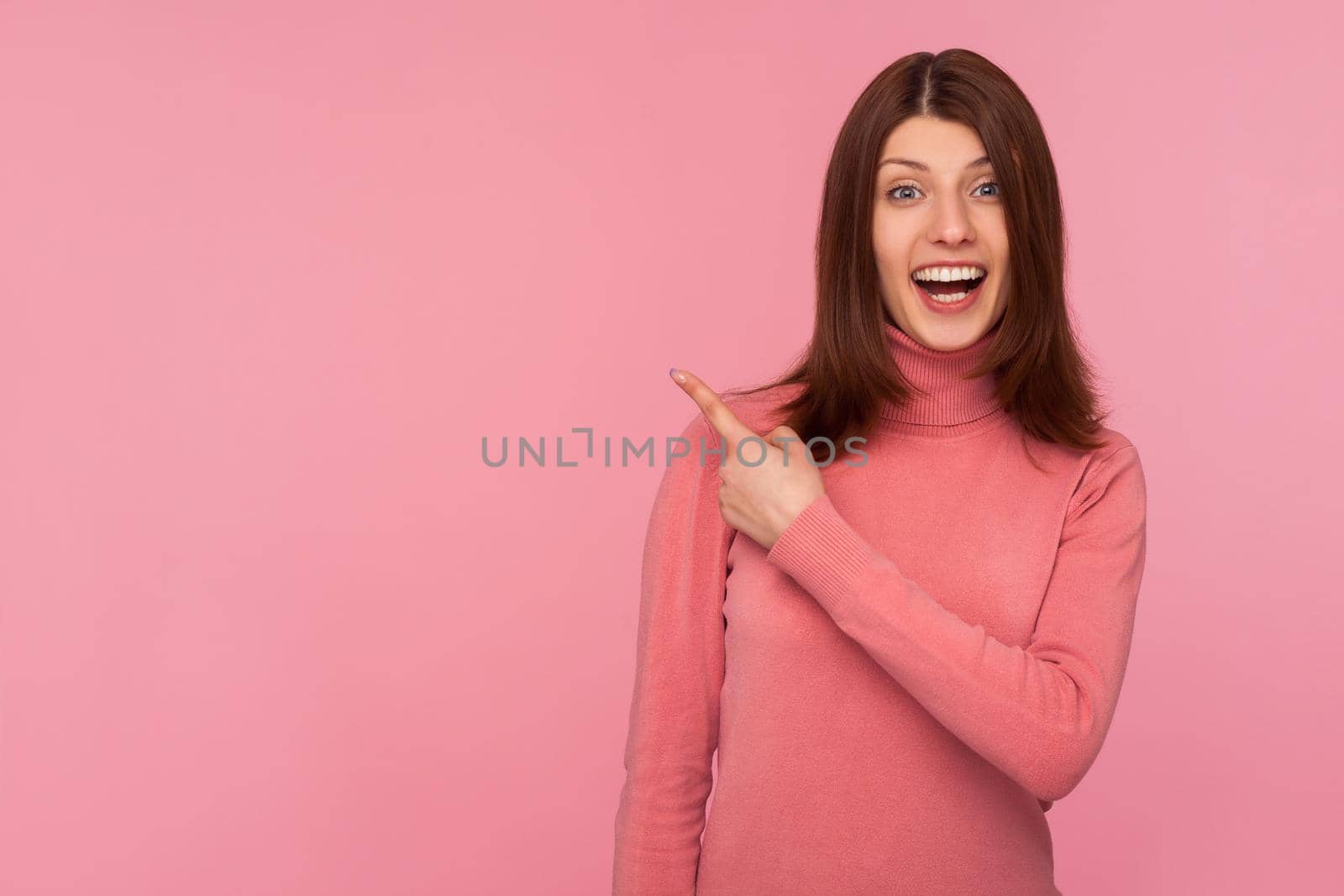 Happy surprised woman in pink sweater pointing finger away with toothy smile, wondering about empty space for your advertisement, template. Indoor studio shot isolated on pink background