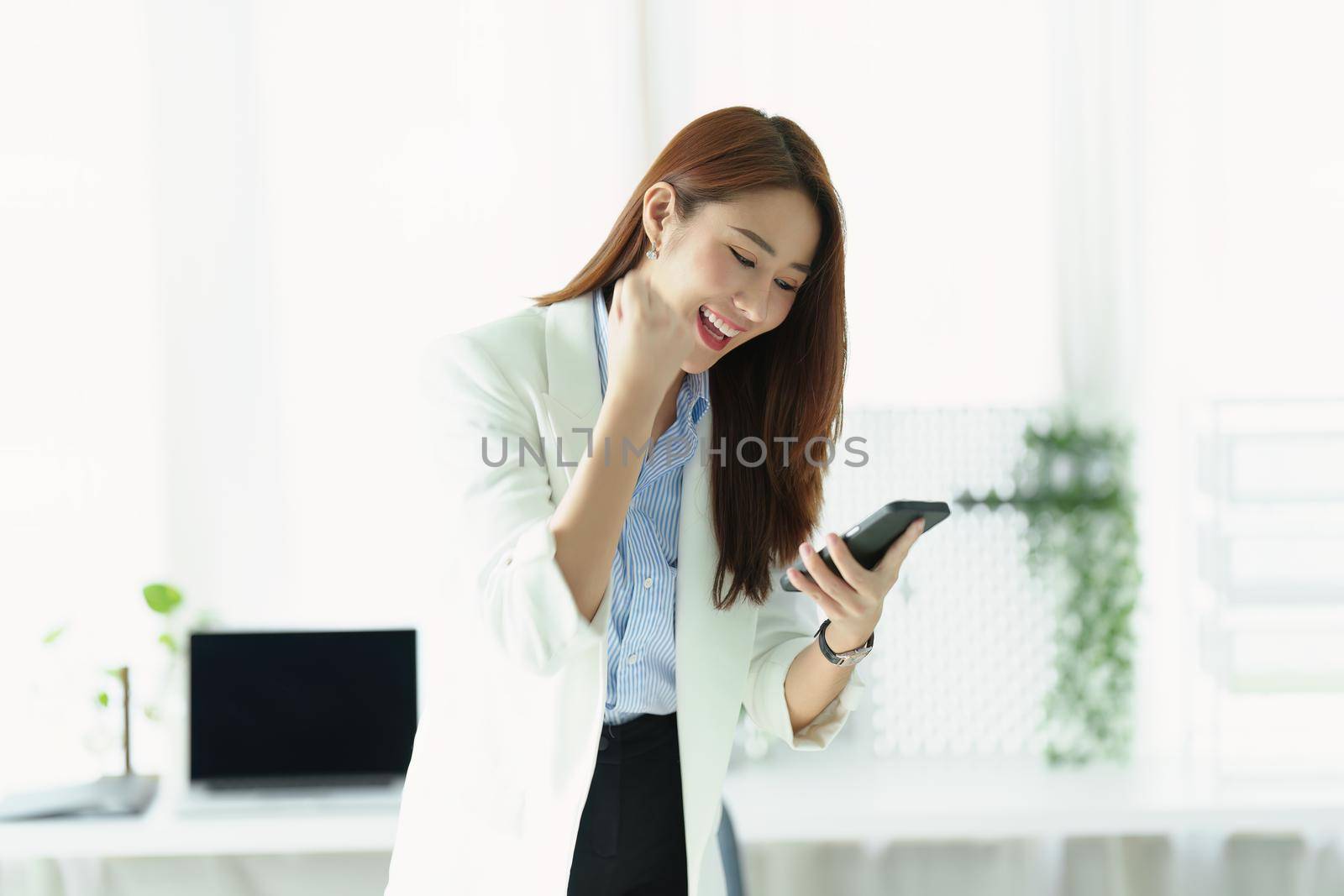 Portrait of a successful Asian businesswoman or business owner expressing excitement and joy using a smartphone mobile in the office.