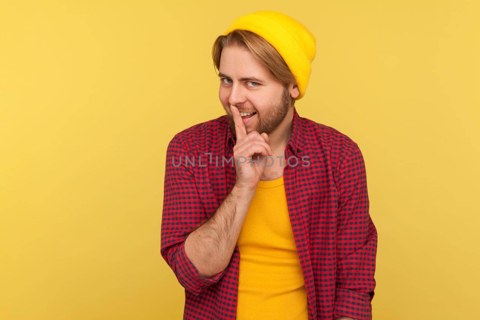 Shh, be quiet. Funny hipster bearded guy in beanie hat and checkered shirt shushing with finger on lips hand gesture, smiling playful mysterious, asking for secrecy. indoor studio shot isolated