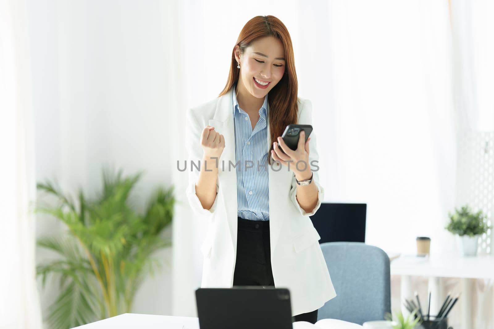 Portrait of a successful Asian businesswoman or business owner expressing excitement and joy using a smartphone mobile in the office by Manastrong
