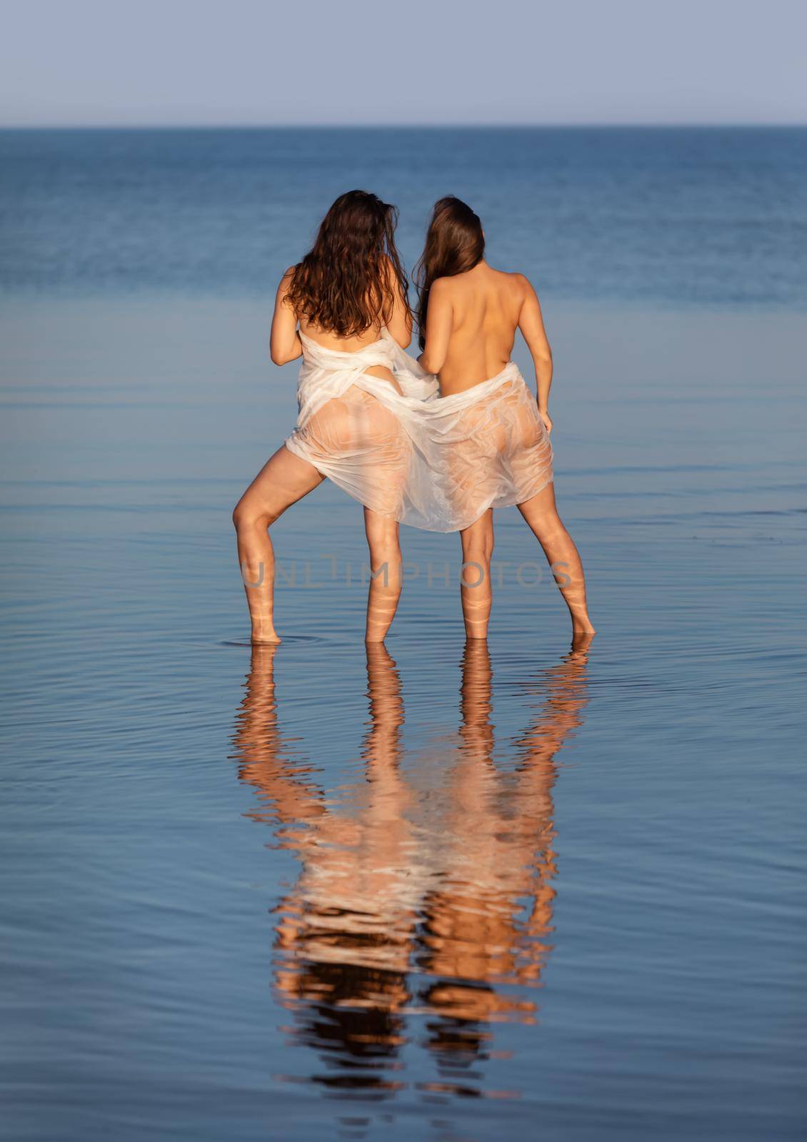 Beautiful naked girl outdoors enjoying nature. Two young nude women wrapped in white shawl posing on sea background. Sexy naked brunettes enjoying summer time outdoors. View from the back