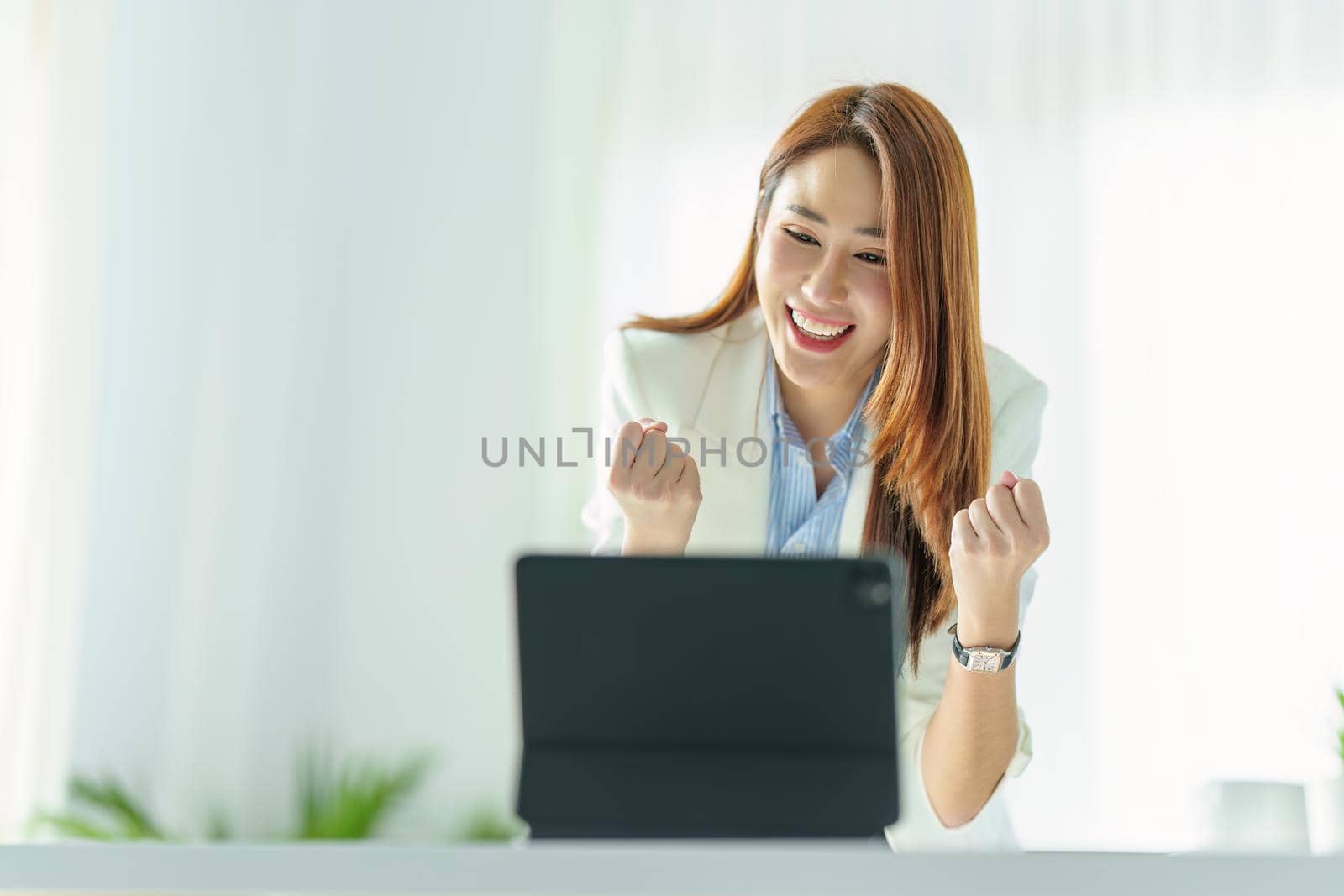 Portrait of a successful Asian businesswoman or business owner expressing excitement and joy using a tablet computer in the office by Manastrong