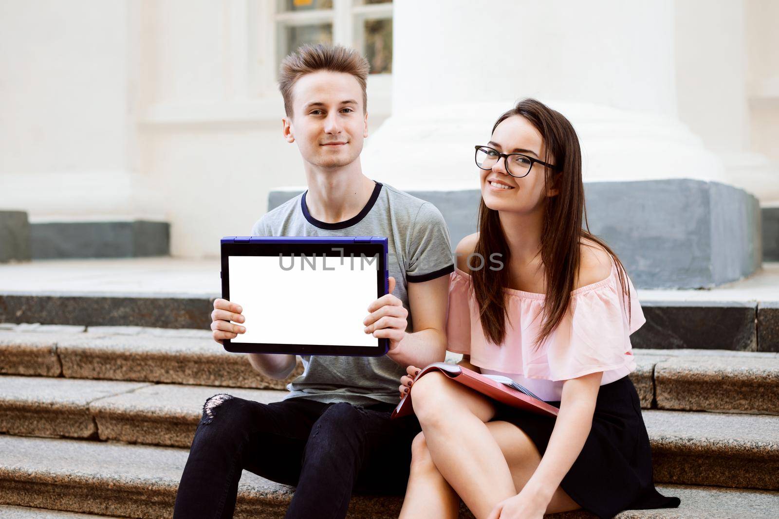 Smiling man and woman using modern laptop convertible computer showing screen to the camera sitting on stairs of old university by VitaliiPetrushenko