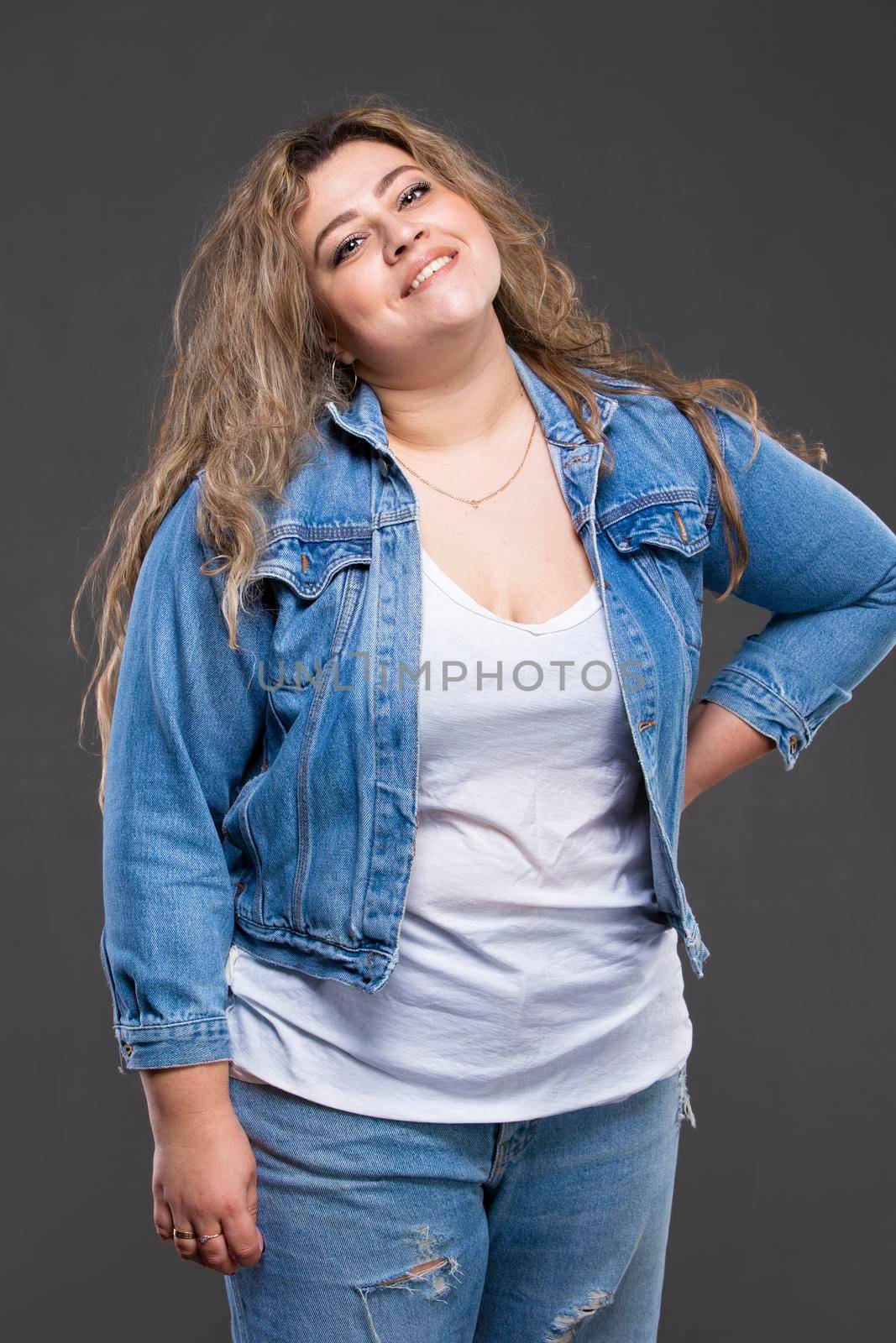 A beautiful fat woman in a denim jacket looks at the camera and smiles on a gray background.