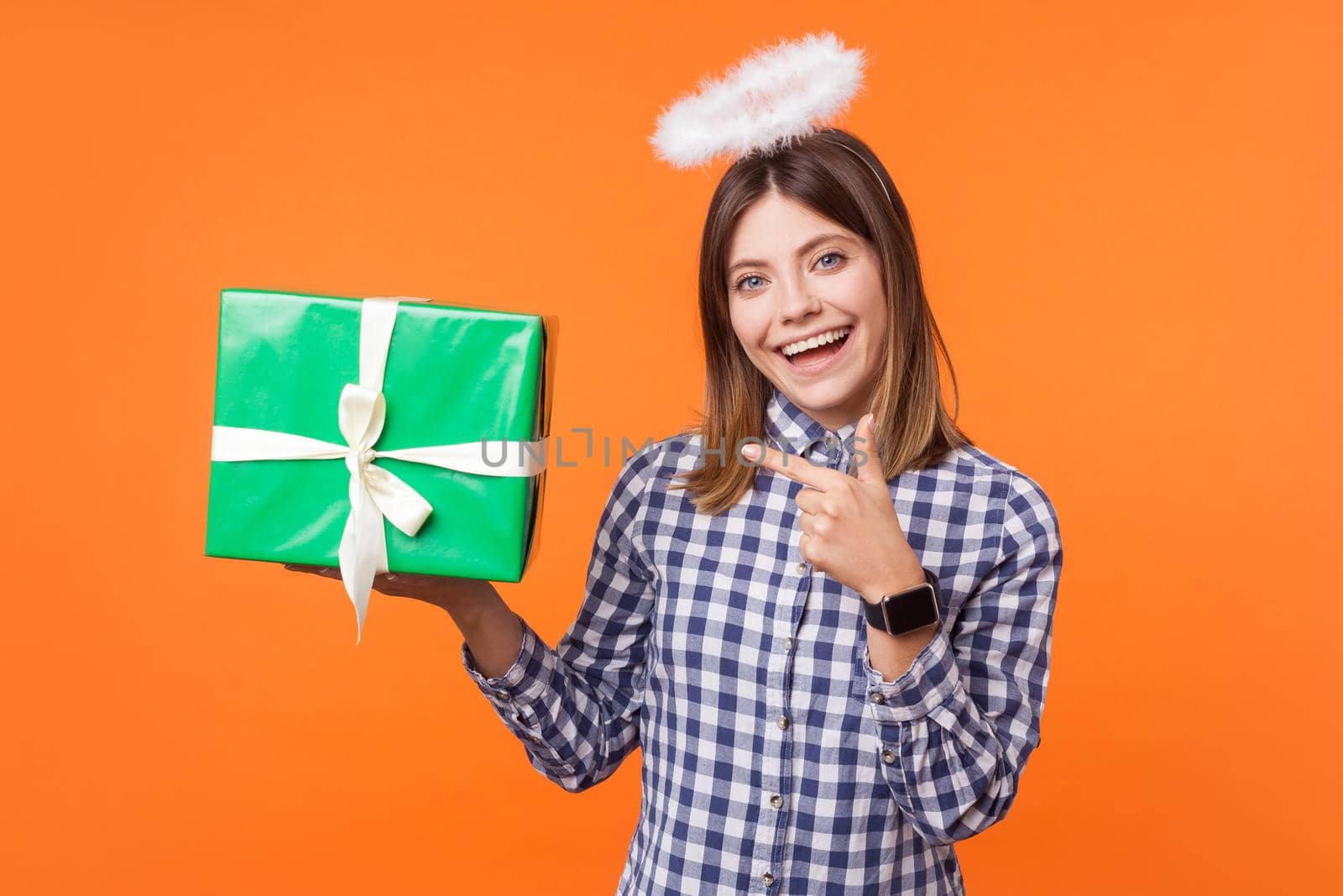 Portrait of angelic positive brunette woman with charming smile wearing checkered shirt standing with halo on head and pointing at gift box, charity. indoor studio shot isolated on orange background