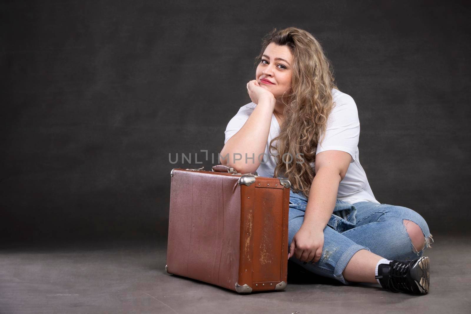Beautiful fat woman in denim clothes with an old retro suitcase on a gray background. Plus size model girl. by Sviatlana