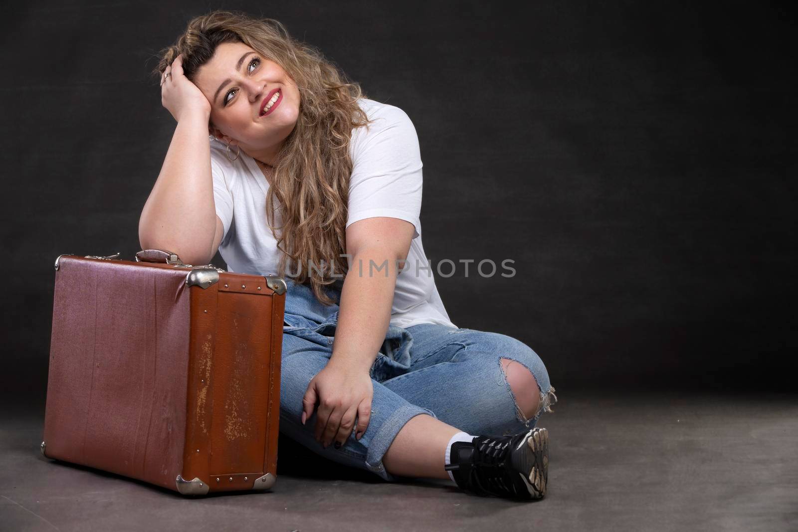 Beautiful fat woman in denim clothes with an old retro suitcase on a gray background. Plus size model girl. by Sviatlana