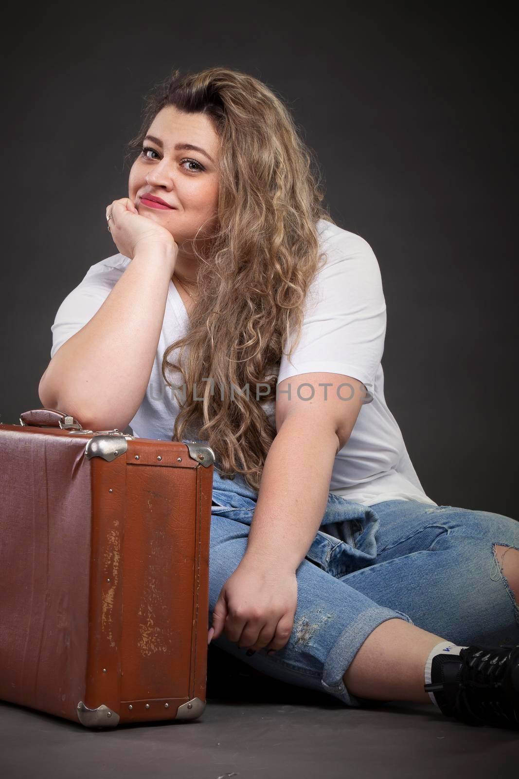 Beautiful fat woman in denim clothes with an old retro suitcase on a gray background. Plus size model girl.