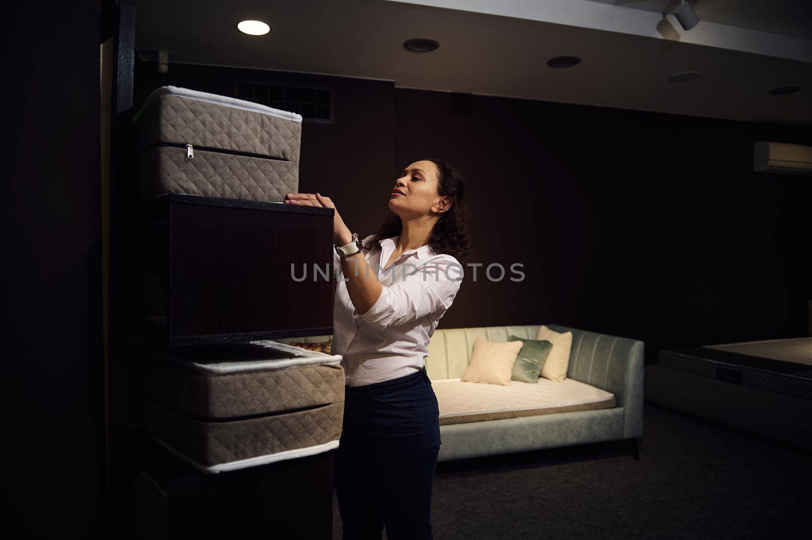 Charming dark-haired multi-ethnic woman, designer, retail consultant, sales manager, retailer, warehouse worker lays out samples of orthopedic mattresses on a stand