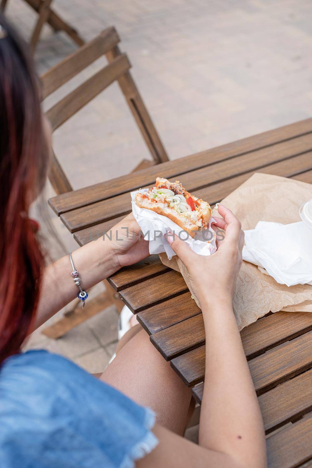 Summer vacation, street food eating. closeup of woman hands holding hamburger, woman eating fast food at street cafe, view from behind