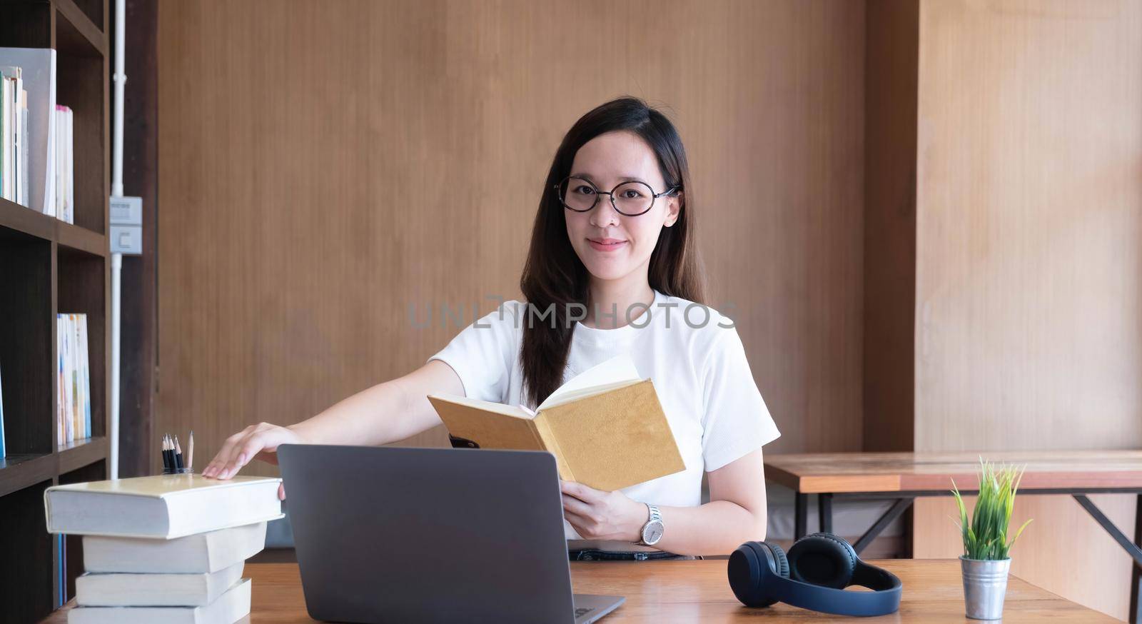 Smiling asian undergraduate teen girl student study in library with laptop books doing online research for coursework, making notes for essay homework assignment, online education e-learning concept by wichayada