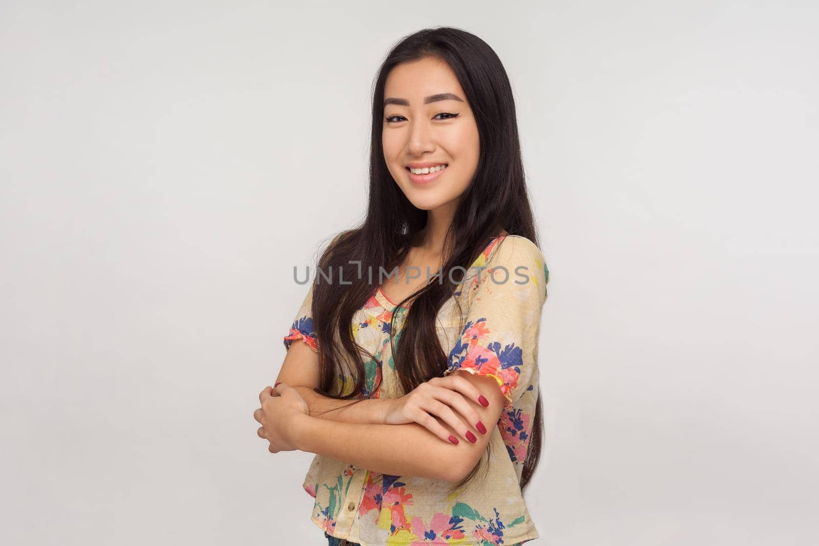 Portrait of calm friendly kind asian girl in summer blouse standing with crossed hands and looking at camera with toothy smile, optimistic outlook. indoor studio shot isolated on gray background