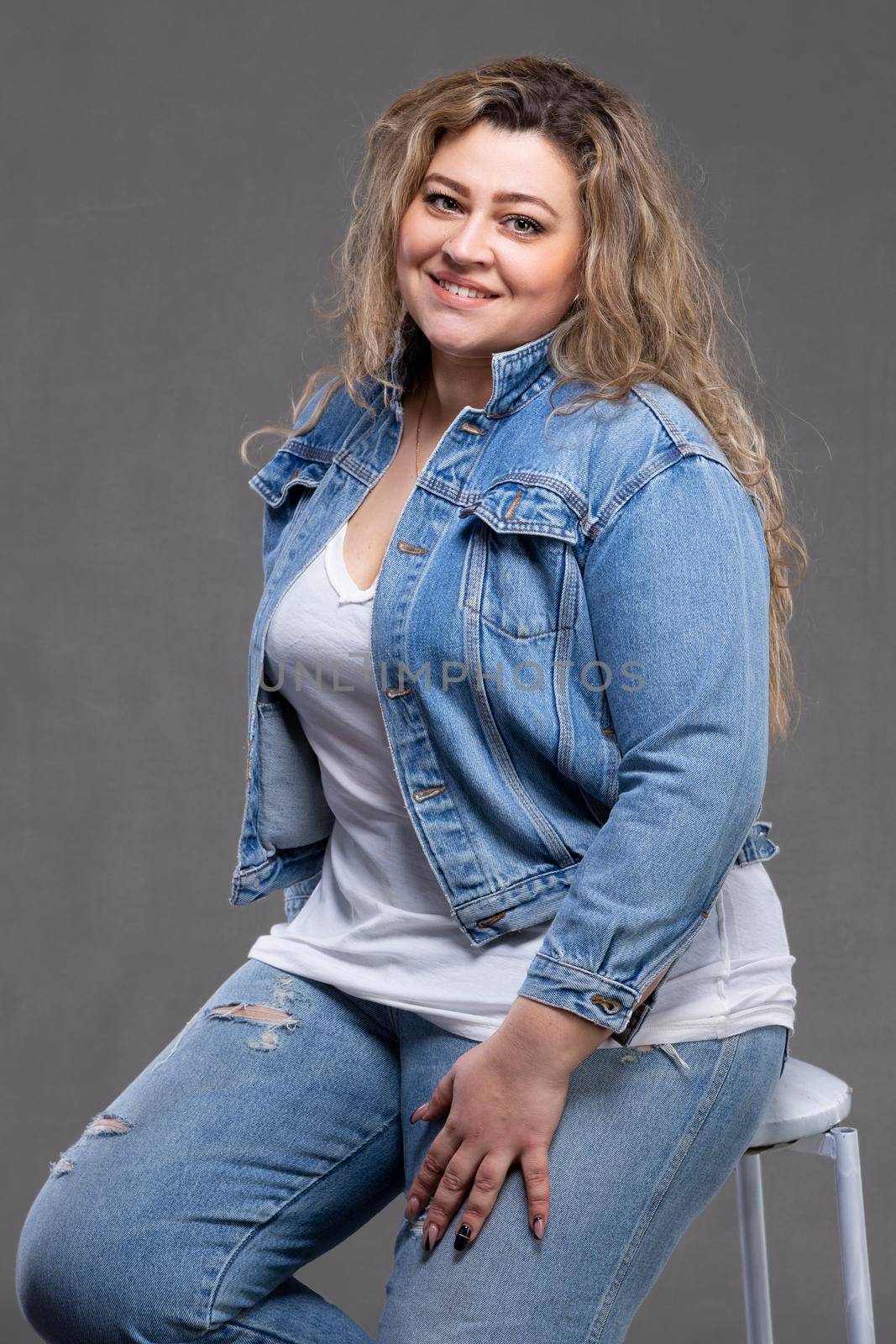 A beautiful fat woman in a denim jacket looks at the camera and smiles on a gray background.