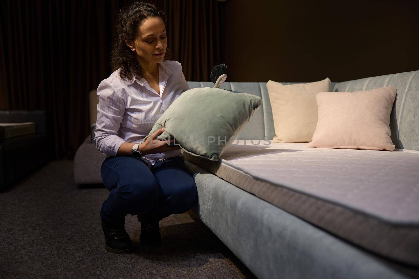 African American sales assistant, consultant in a designer furniture store, straightens a green velour pillow and dusts off a comfortable modern new upholstered sofa bed with orthopedic mattress