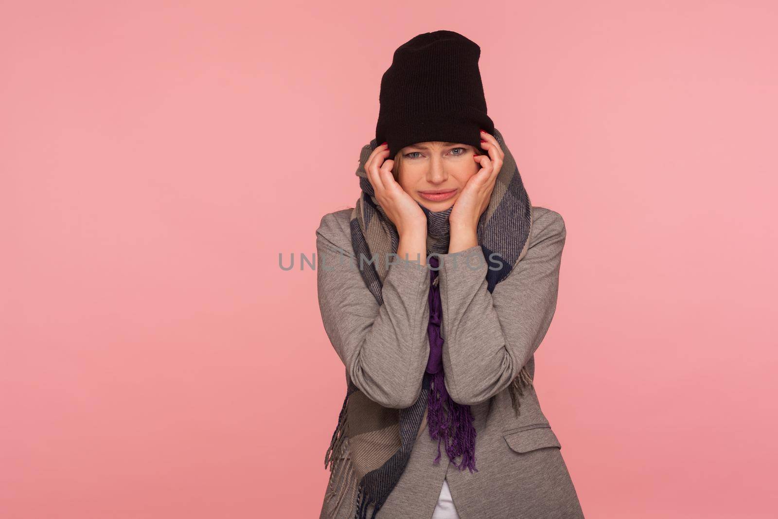 Portrait of depressed woman in warm hat and scarf holding her head suffering migraine and fever, first flu symptoms, looking with frustrated dismal expression. studio shot isolated on pink background