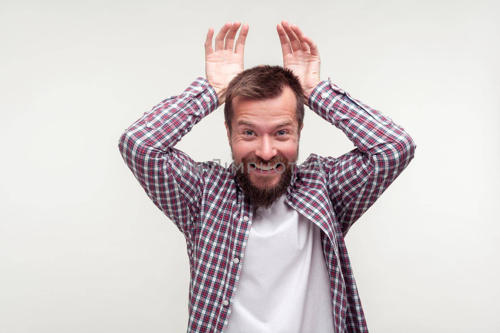 Portrait of funny happy bearded man in plaid shirt making bunny ears gesture, smiling joyfully at camera and feeling childish carefree, optimistic view. indoor studio shot isolated on white background