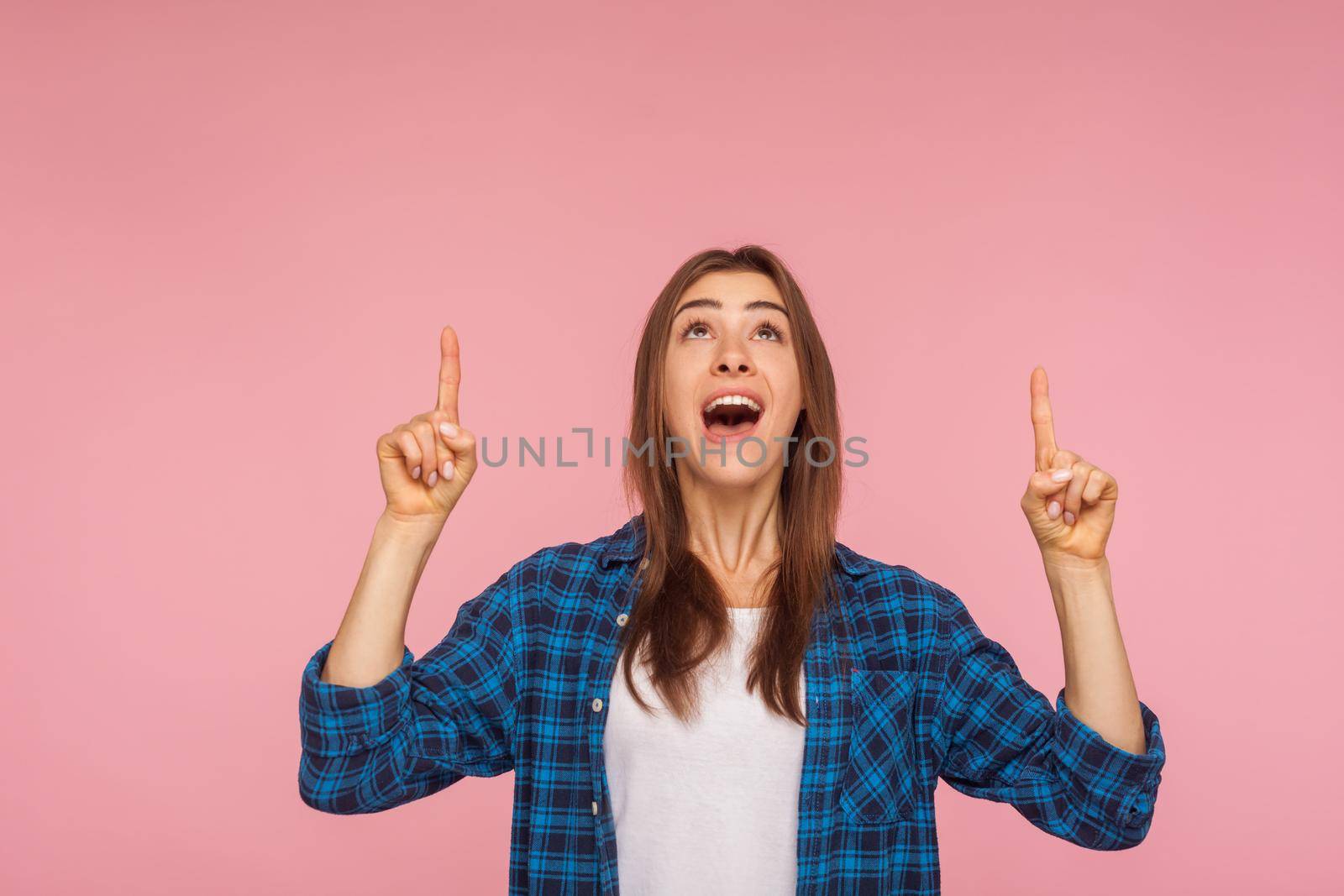Wow, look above, crazy advertisement. Portrait of amazed brunette girl in checkered shirt pointing up at empty place for ad content and expressing surprise. studio shot isolated on pink background
