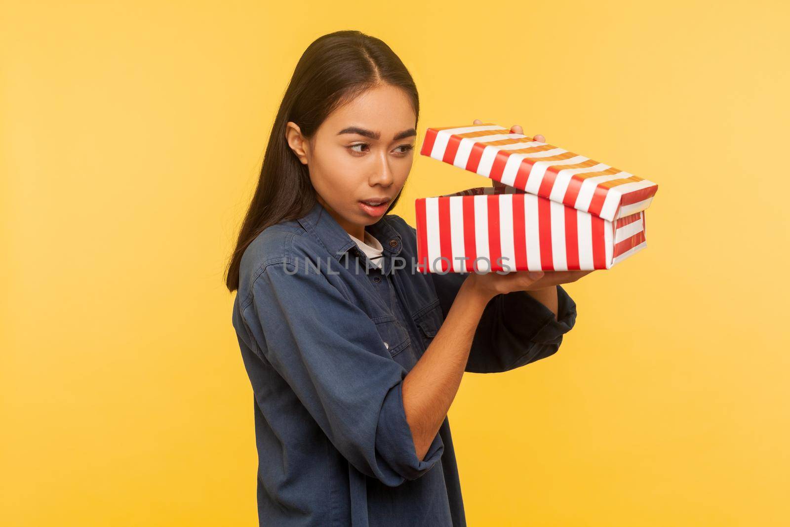 Portrait of curious funny girl in denim shirt opening gift and looking into box with interest, peeking inside in anticipation of nice present surprise. indoor studio shot isolated on yellow background