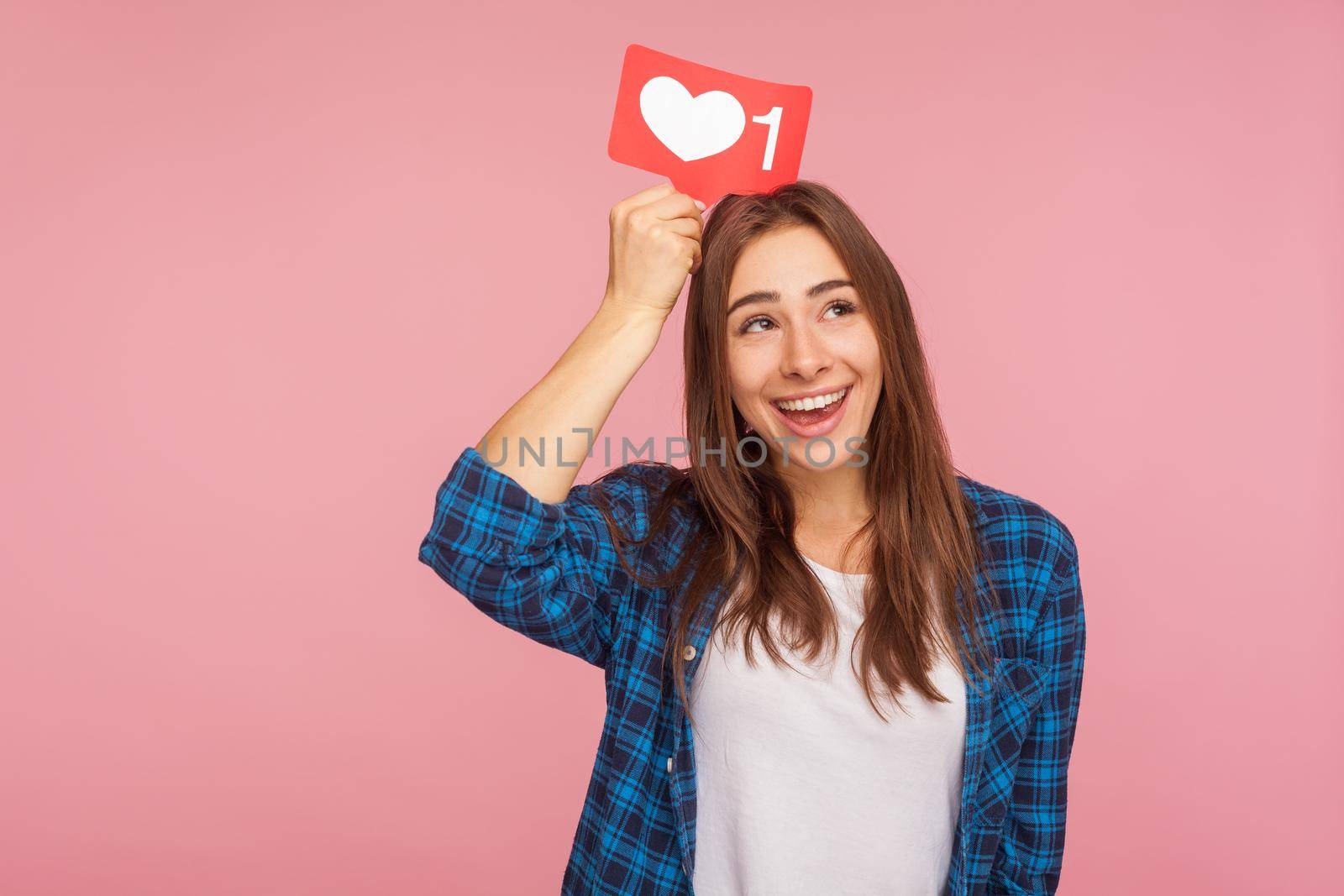 Portrait of beautiful positive blogger girl in checkered shirt dreaming of more likes in social media, holding heart icon on head, thinking over trendy interesting content. studio shot pink background