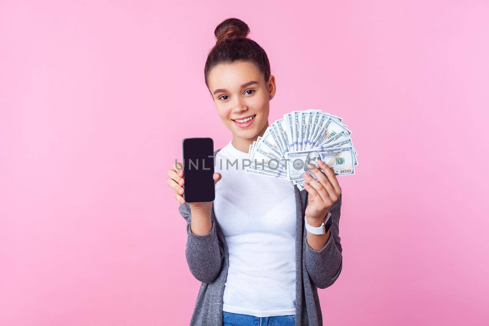 Mobile banking apps. Portrait of beautiful brunette teen girl with bun hairstyle in casual clothes showing cell phone and dollars, smiling at camera. indoor studio shot isolated on pink background