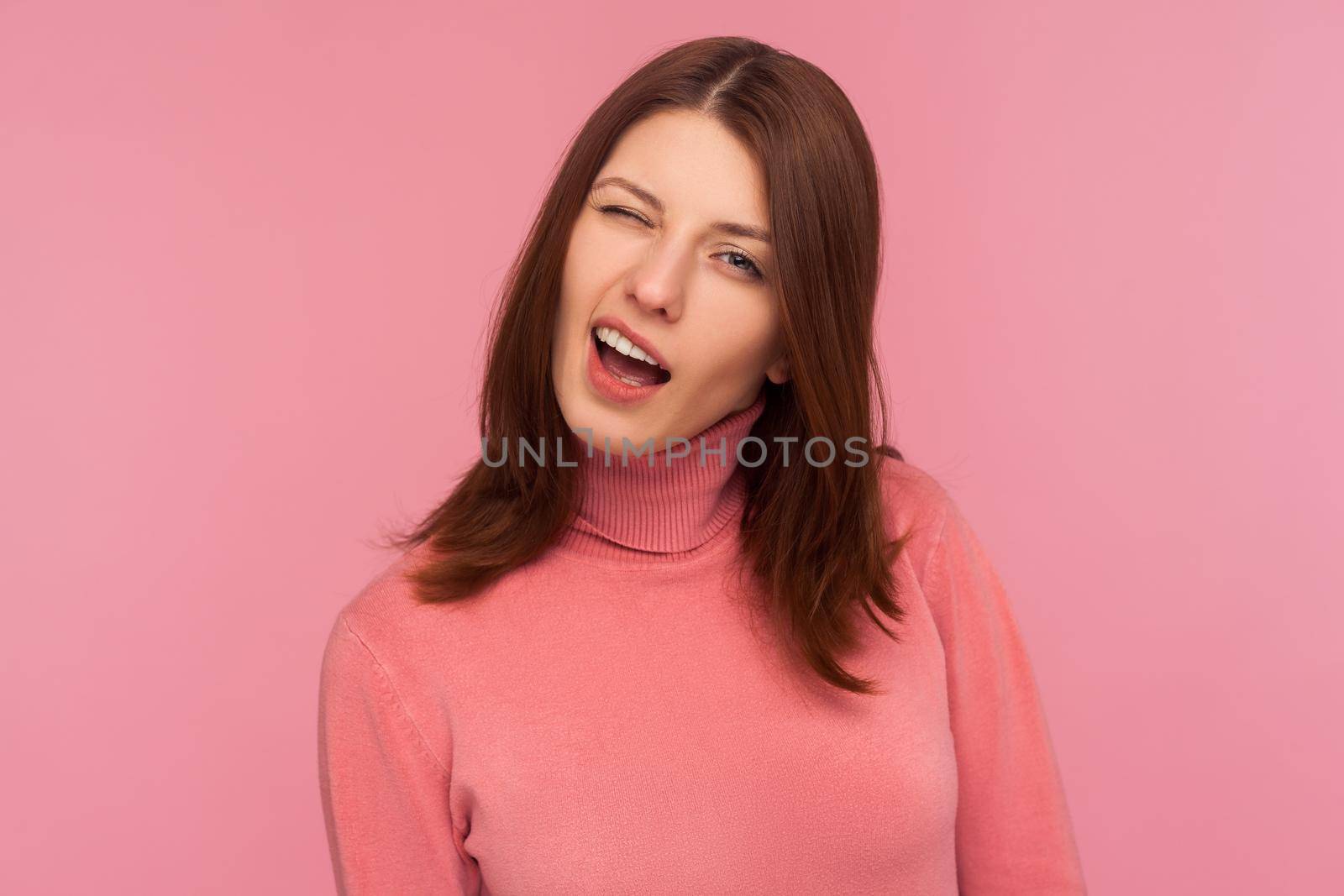 Closeup portrait of coquettish funny brunette woman in pink sweater winking looking at camera, cheering up with wink. Indoor studio shot isolated on pink background