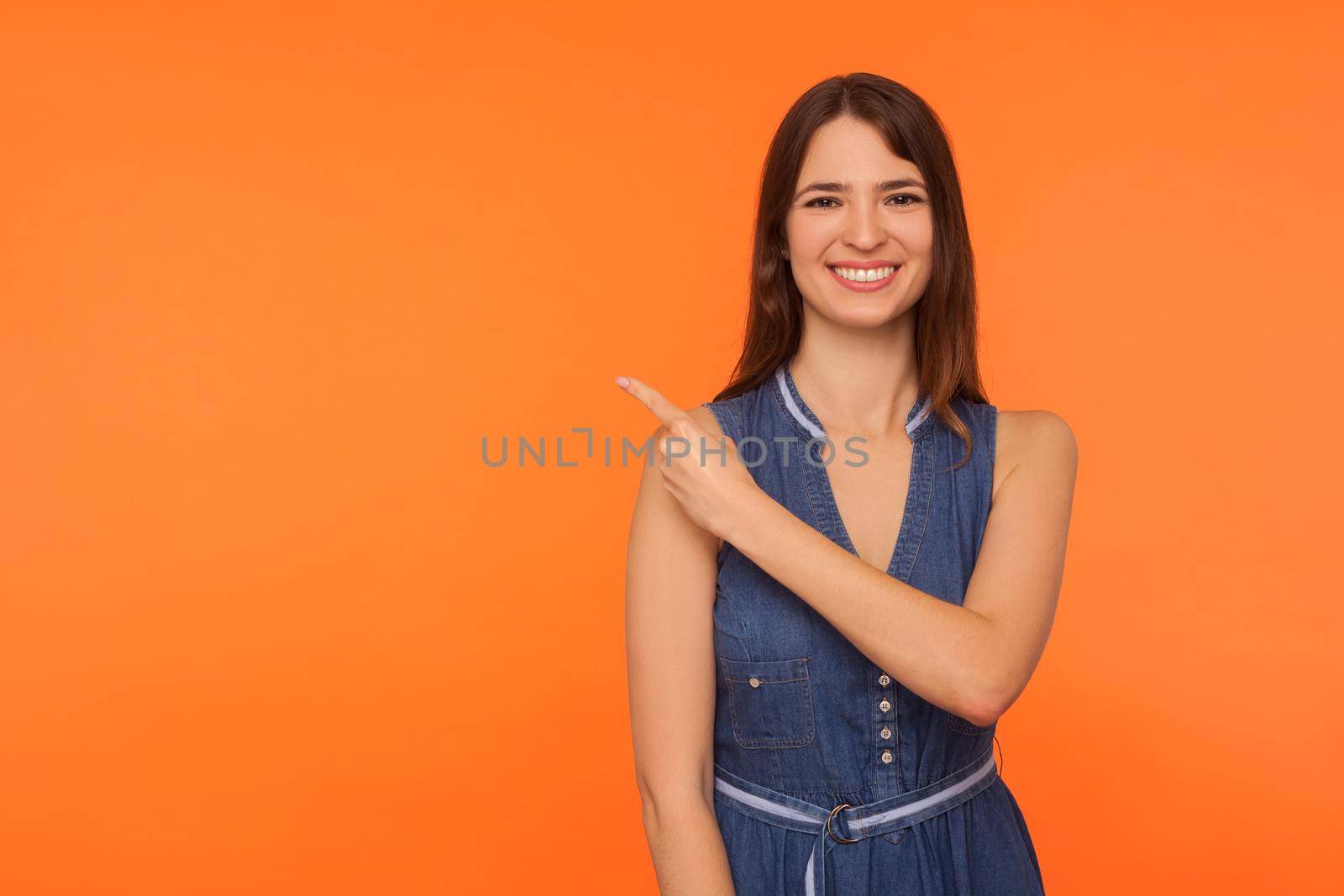 Look, advertising area. Cheerful happy brunette woman in denim dress smiling and pointing finger to empty place on orange background, showing copy space for promotional text or image. studio shot