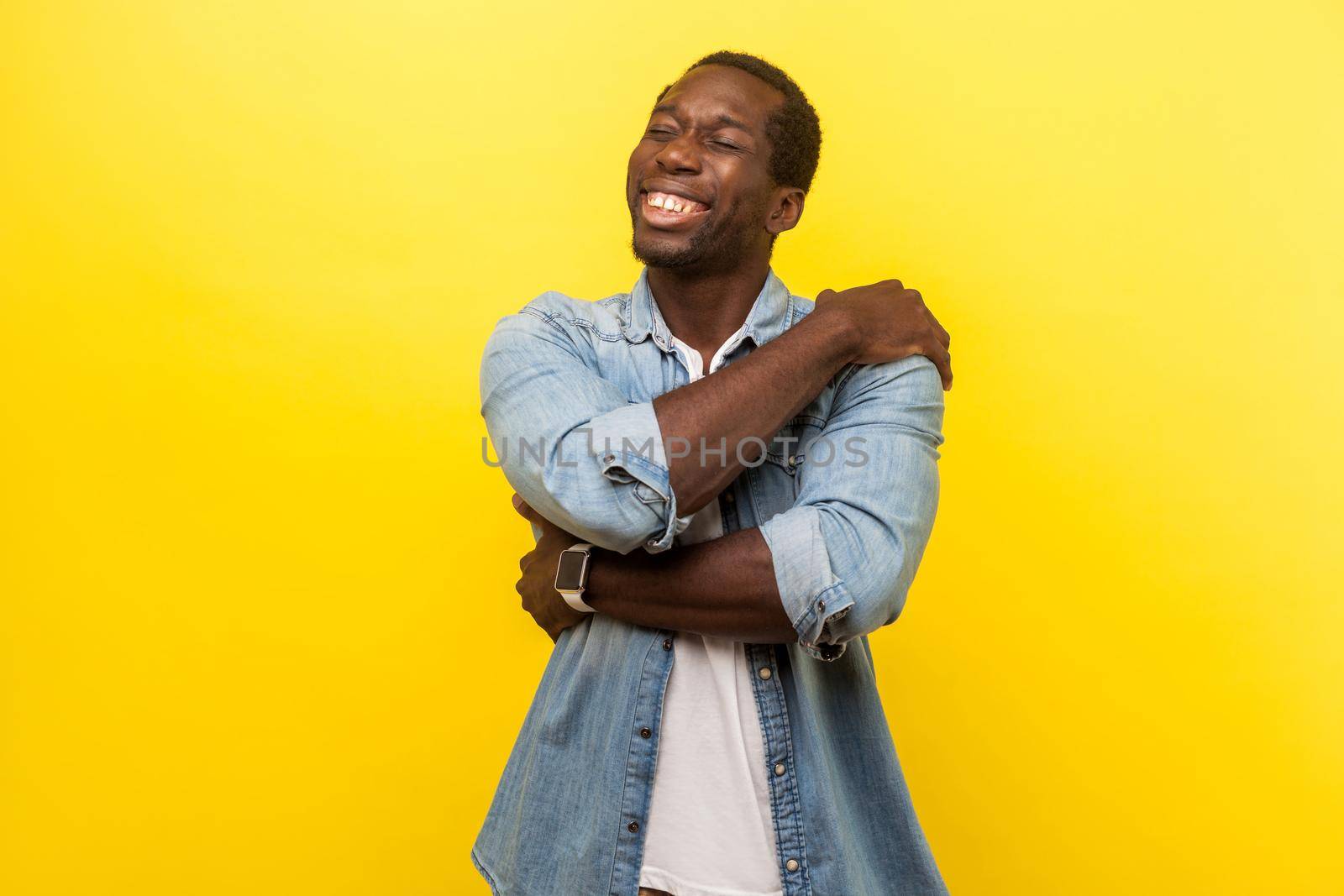 I love myself. Portrait of self-satisfied happy man in denim casual shirt cuddling himself and smiling with pleased delighted expression, egoist. indoor studio shot isolated on yellow background