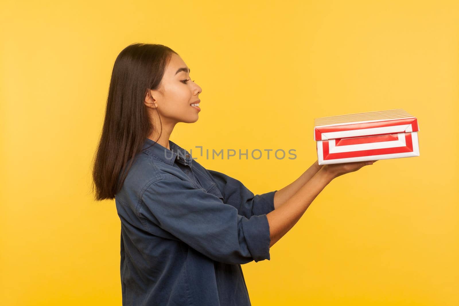 Take this present. Side view of generous happy brunette girl in denim shirt giving box and smiling, sharing holiday gift, congratulating on birthday. indoor studio shot isolated on yellow background
