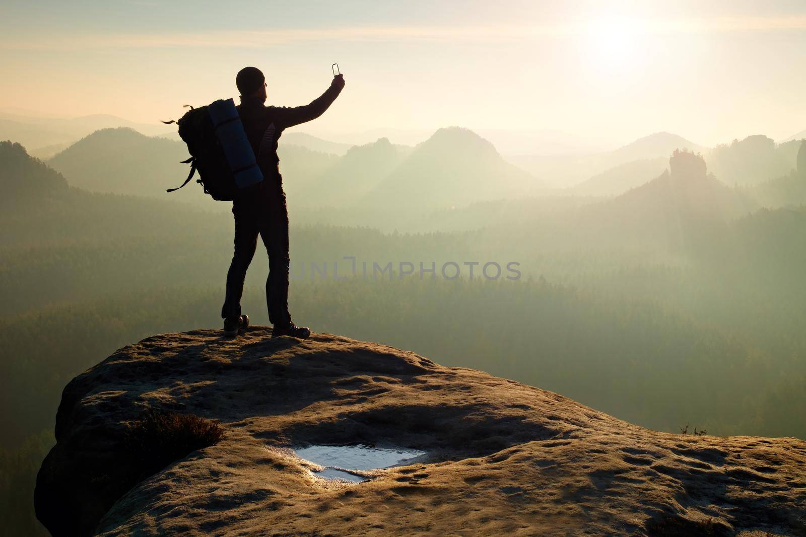 Tourist with backpack takes photos with smart phone on the rocky peak. Dreamy fogy valley below