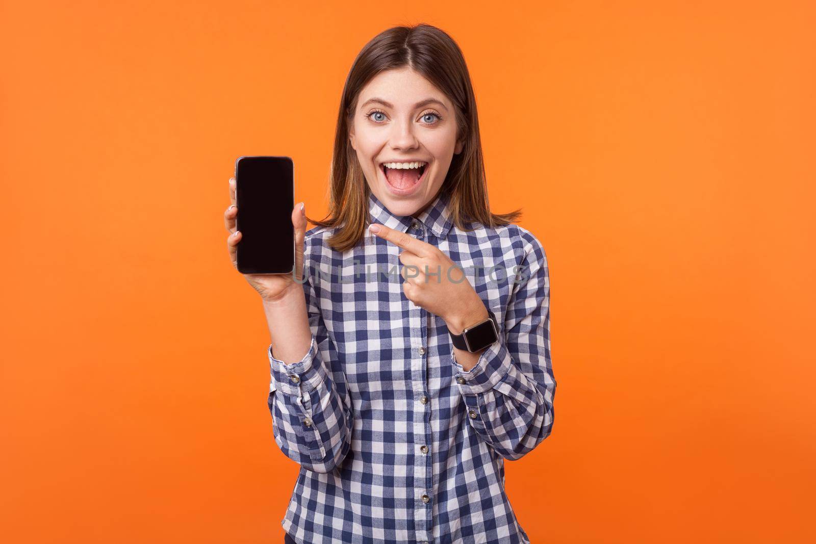 Portrait of shocked attractive brunette woman with charming smile wearing checkered shirt standing pointing at phone, amazed and wow facial expression. indoor studio shot isolated on orange background
