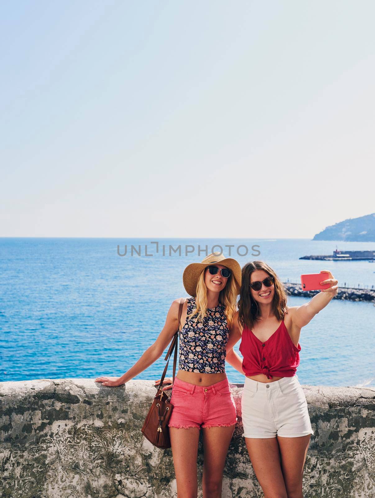 Two friends taking selfies on summer vacation. Girl friends on travel adventure taking photos for social media with smartphone by YuriArcurs