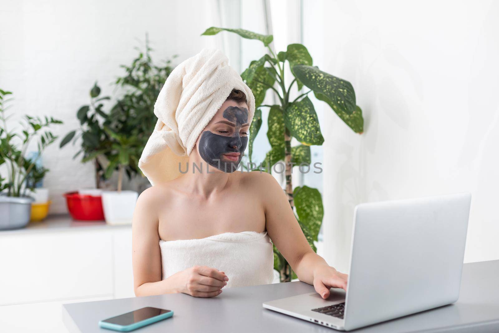 Beautiful young woman working on laptop computer while sitting at the living room with face mask. Beautiful young woman smiling with face mask and looking at laptop screen.