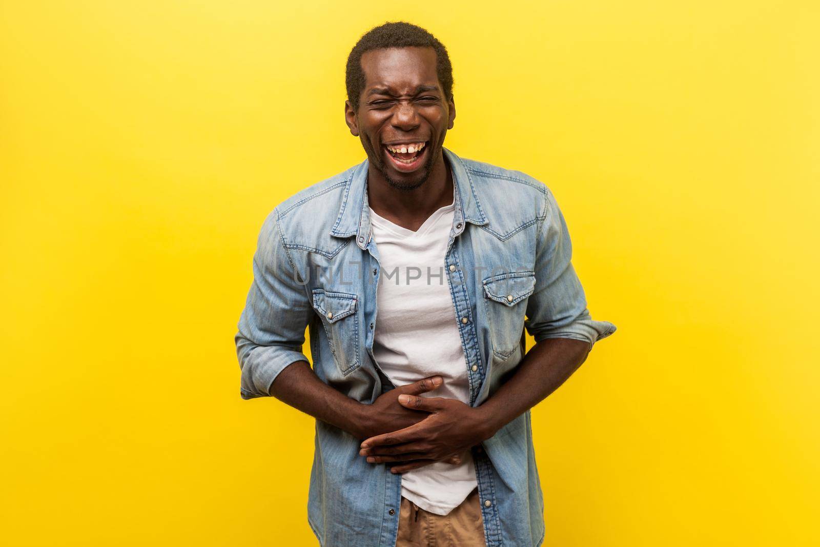 Portrait of overjoyed happy man in denim casual shirt with rolled up sleeves laughing hard and holding hands on stomach, feeling amused by joke. indoor studio shot isolated on yellow background
