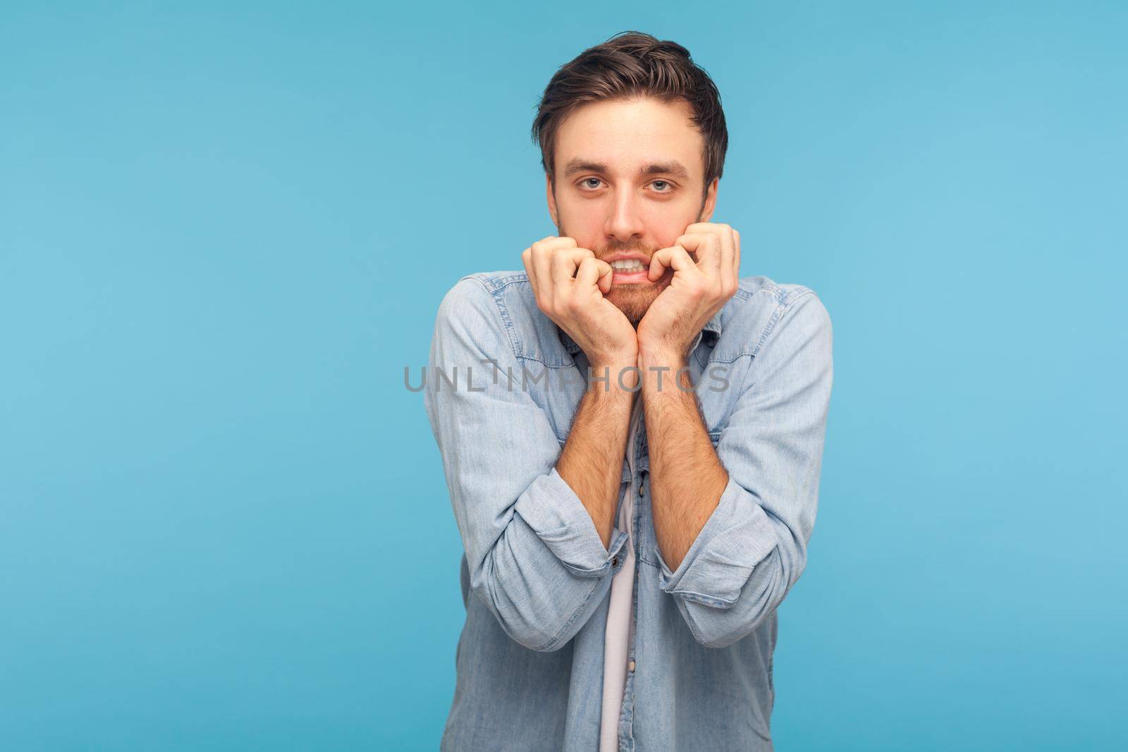 Anxiety disorder, depression. Portrait of stressed out, worried man in worker denim shirt biting nails, nervous about troubles, panicking and looking scared. studio shot isolated on blue background