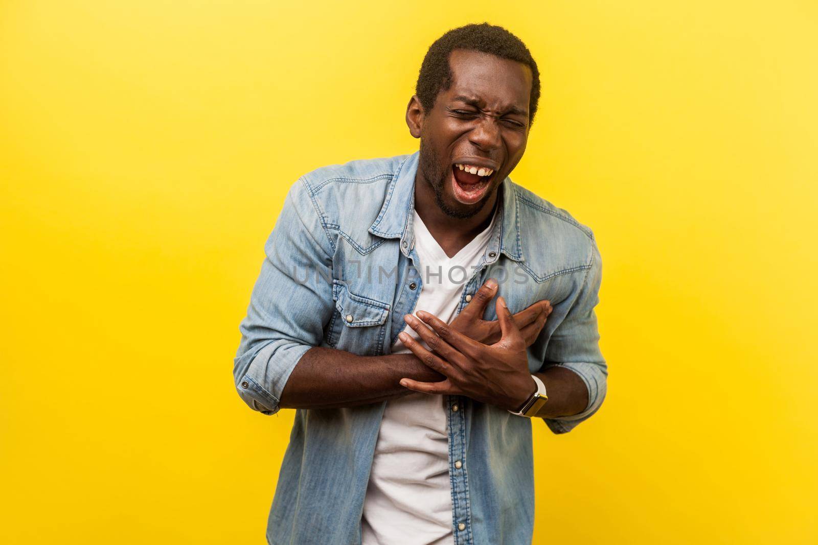 Portrait of ill stressed man in denim casual shirt shouting and grimacing from unbearable pain, clutching chest, having heart attack or painful cramps. indoor studio shot isolated on yellow background